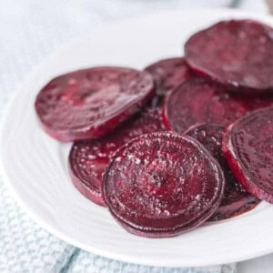 A white plate holds several slices of roasted beets, arranged in overlapping layers. The deep purple, slightly glossy beets contrast beautifully against the light, textured background.
