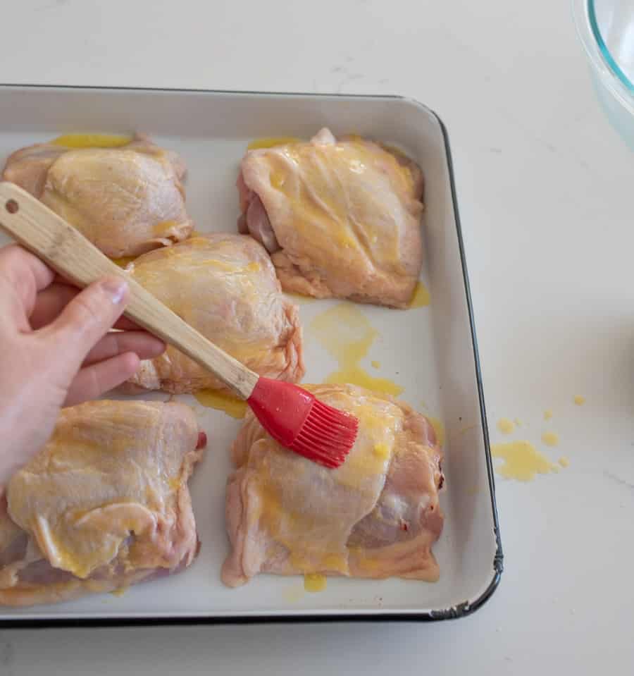 woman holding red silicone basting brush brushing oil on chicken thighs on white baking sheet