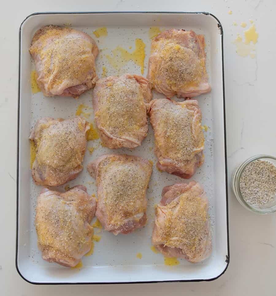 uncooked chicken thighs with seasoning on baking sheet next to clear bowl of season salt on white countertop