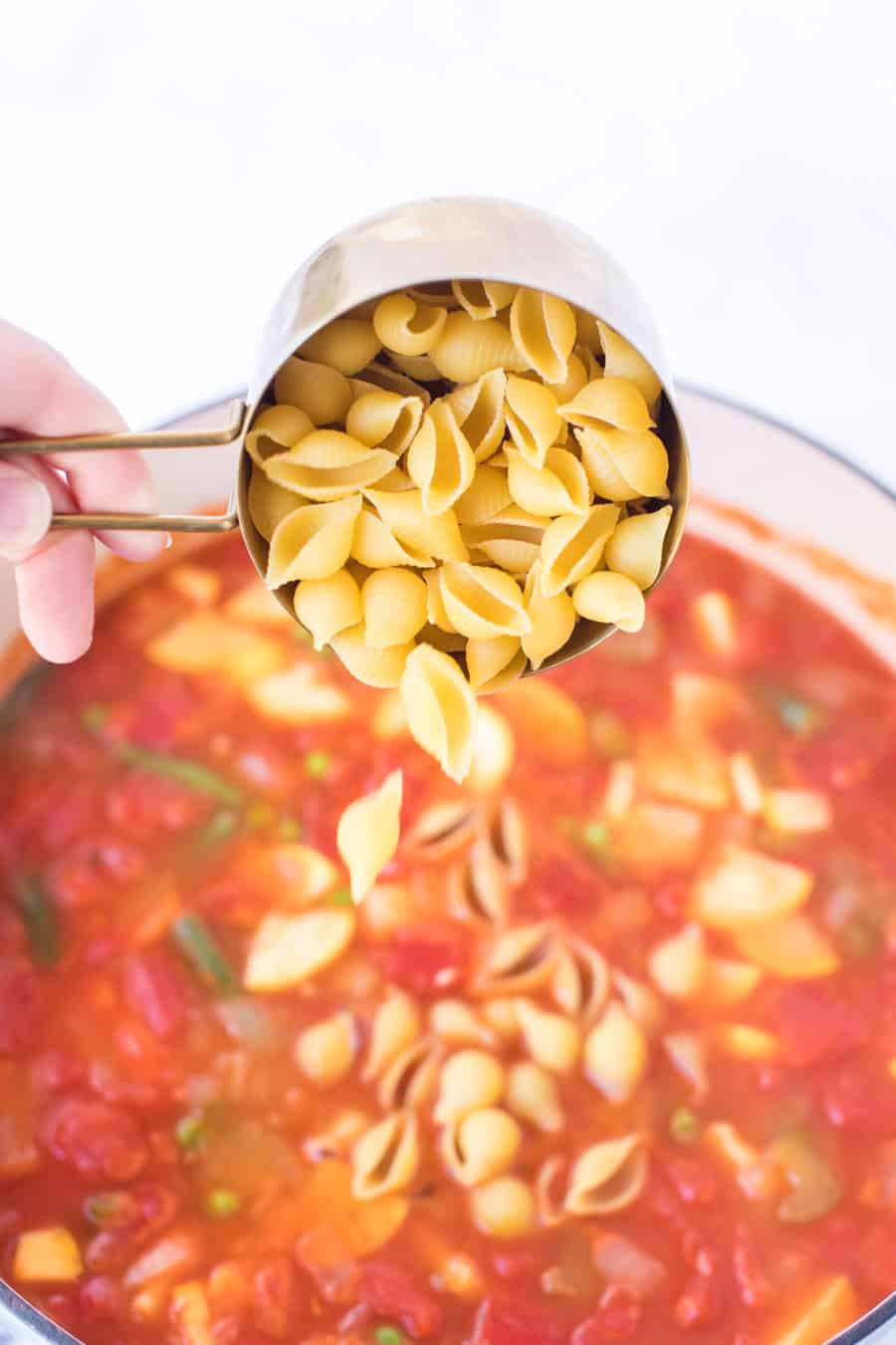 cup of pasta shells being poured into pot of minestrone soup