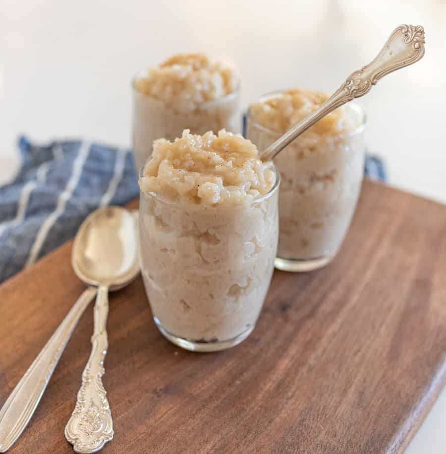 3 glasses of rice pudding with spoon on wooden cutting board and blue and white striped towel