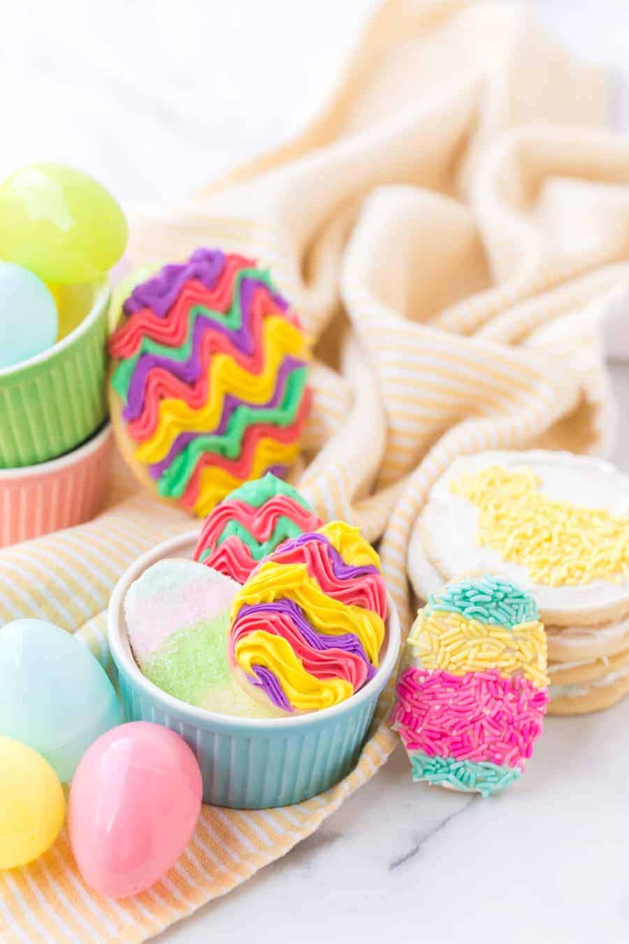 long image of iced sugar cookies and cookies with sprinkles in ramekins with plastic easter eggs on yellow and white striped towel