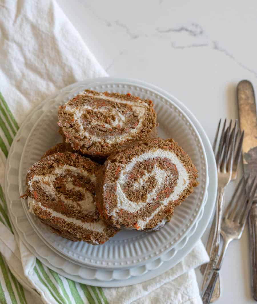 three slices of carrot cake roll on round white plate stack and green striped towel with forks and knife