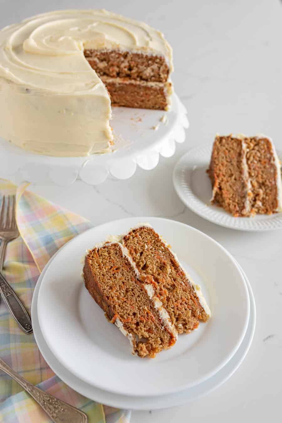 carrot cake on white cake stand next to slices of carrot cake on two round white plates with plaid pastel napkin