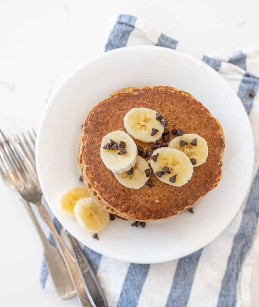 closeup of stack of healthy banana pancakes with banana slices and chocolate chips with blue and white towel and forks on white countertop