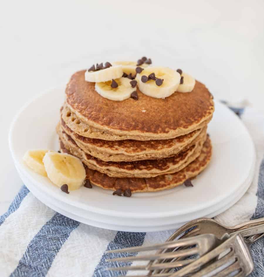 stack of banana pancakes with banana slices and chocolate chips on stack of white plates with forks