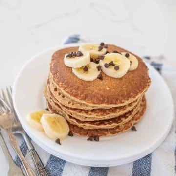 stack of banana pancakes on round white plate with banana slices on top of blue striped towel