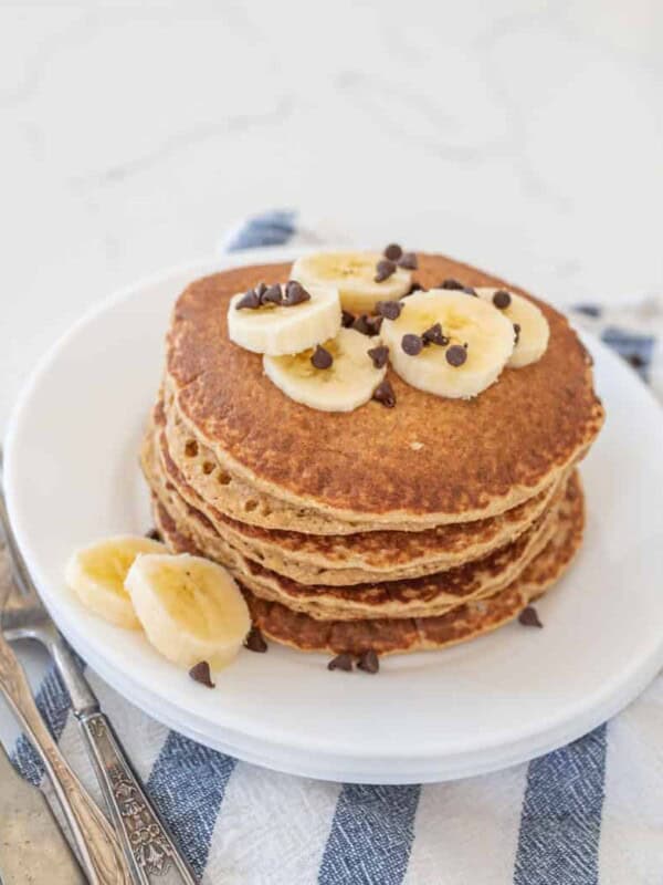 stack of banana pancakes on round white plate with banana slices on top of blue striped towel