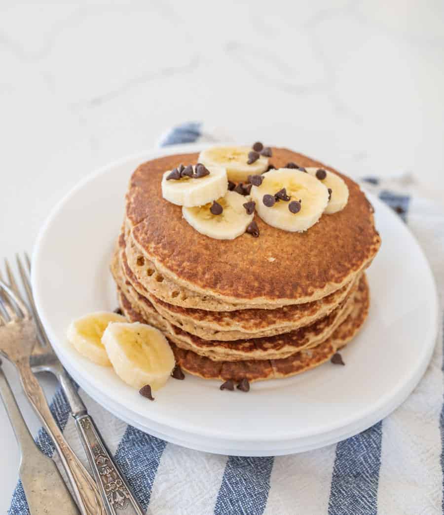 stack of banana pancakes on round white plate with banana slices on top of blue striped towel