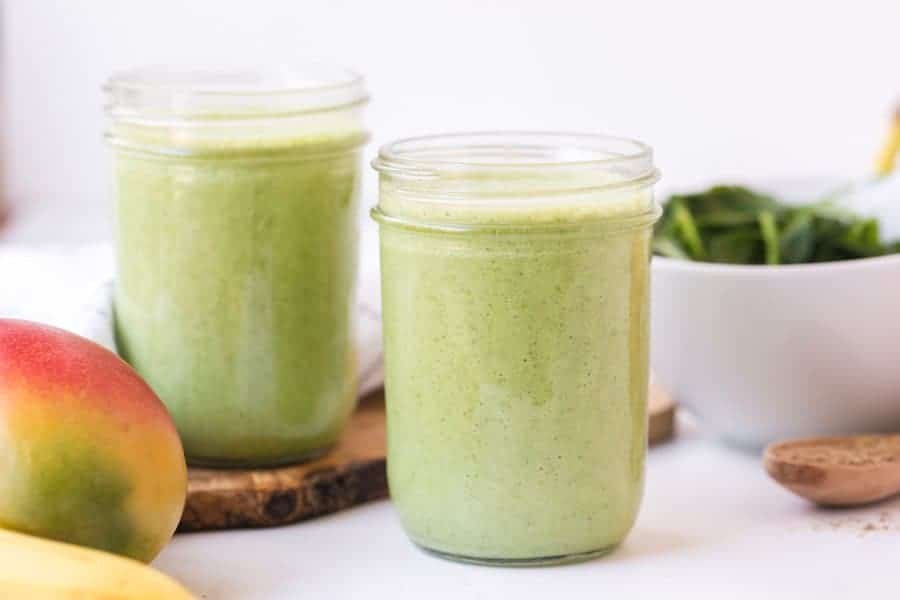 two green smoothies in mason jars next to bowl of spinach and a peach