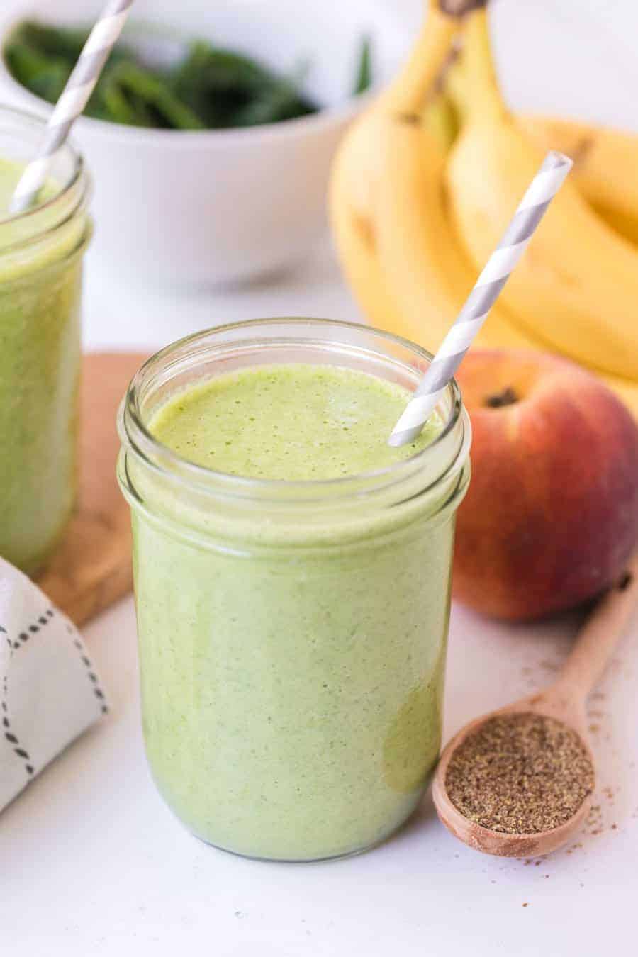 healthy green smoothie is clear jar with a gray and white paper straw next to spinah apple cinnamon and bananas