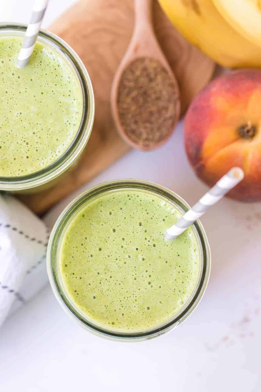 overhead shot of healthy green smoothies with gray and white paper straws on white countertop with peaches and bananas