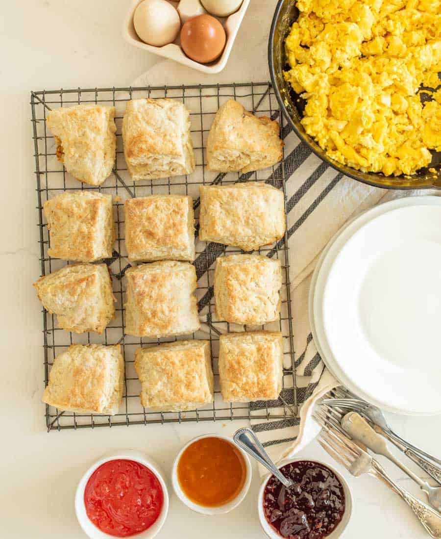 sourdough biscuits on cooking rack next to scrambled eggs jams and jellies and round white plates with utensils