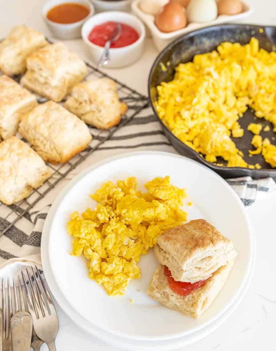 round white plate with scrambled eggs and a sourdough biscuit with jelly next to biscuits on cooling rack and forks