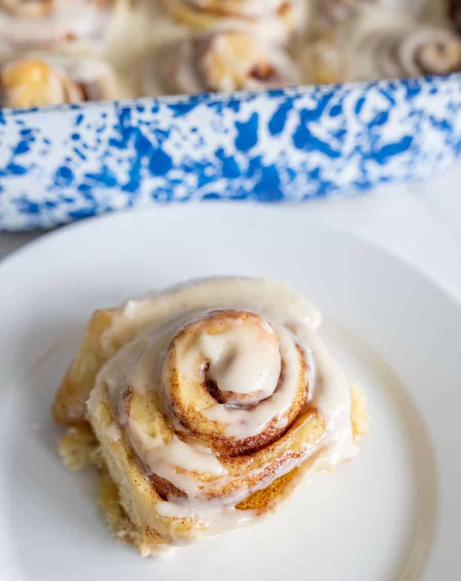 one sourdough cinnamon roll with buttercream icing on round white plate next to blue and white baking dish with rolls