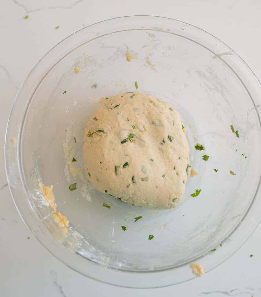 A glass bowl containing a ball of dough with visible green herb pieces, resting on a white countertop.