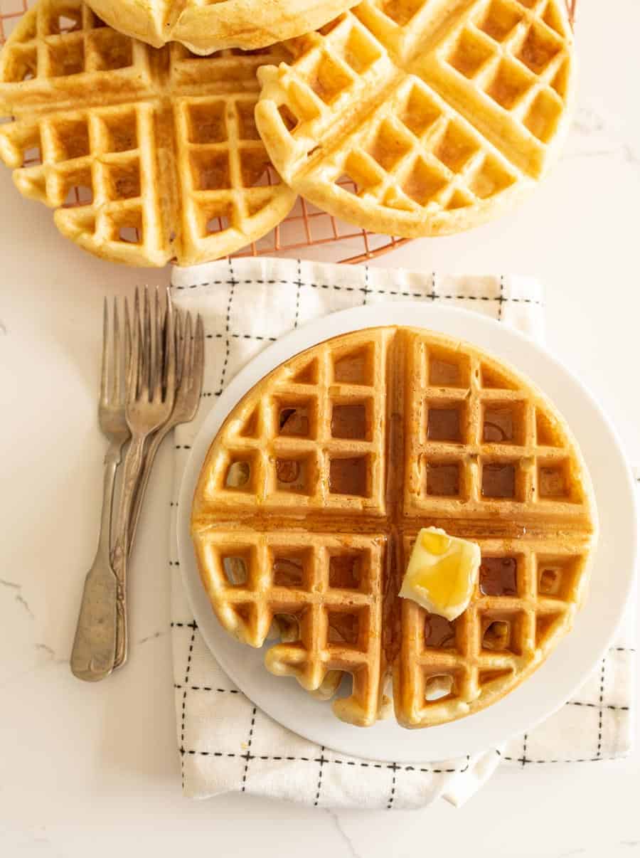 sourdough waffle on round white plate with pat of butter on white towel with forks
