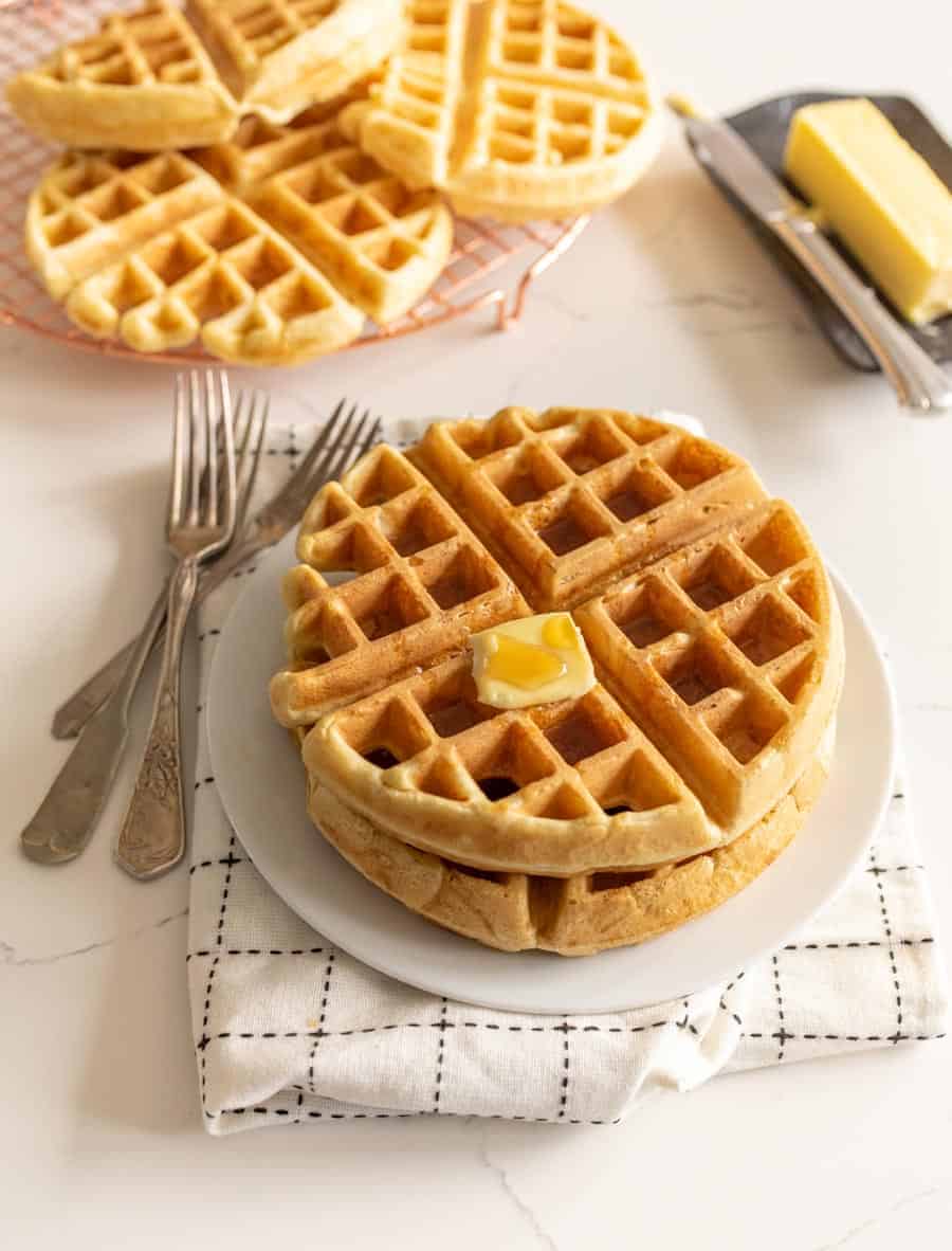 two sourdough waffles with butter on white plates with forks next to stick of butter and waffles on cooling rack