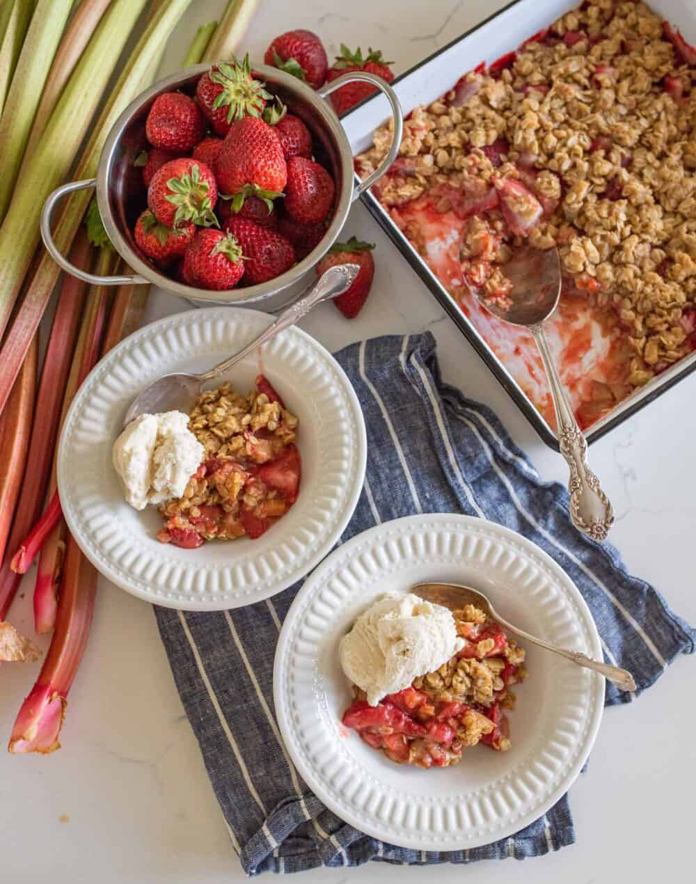 two bowls of dished crisp with ice cream on top next to pan of baked strawberry rhubarb crisp