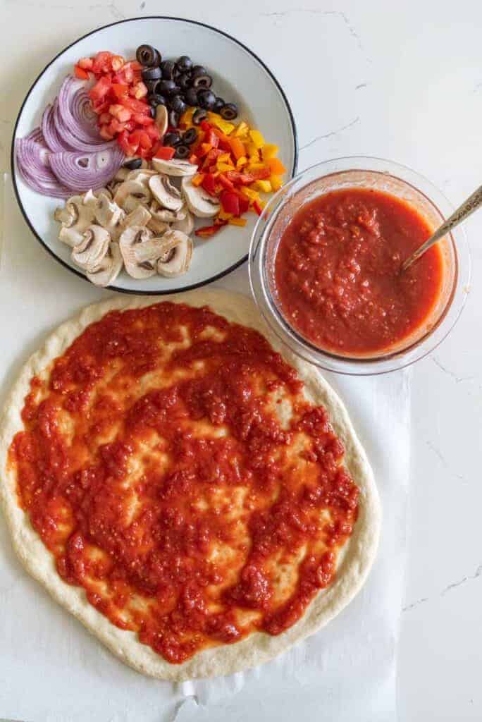 A pizza crust spread with tomato sauce sits on a white surface, ready for a delicious veggie pizza recipe. Nearby are a bowl of sauce with a spoon and a plate with sliced onions, black olives, diced tomatoes, bell peppers, and mushrooms.