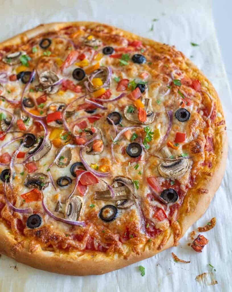 A baked veggie pizza recipe topped with melted cheese, sliced black olives, red onion, mushrooms, diced tomatoes, and chopped herbs, resting on a sheet of parchment paper.