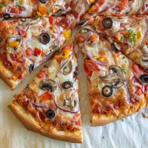 A sliced veggie pizza recipe topped with olives, mushrooms, red onions, tomatoes, and cheese, resting on parchment paper on a light surface.