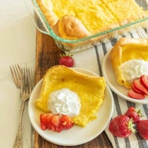 A plate with a serving of Dutch baby, also known as a German pancake recipe, is topped with whipped cream and sliced strawberries. It sits on a cutting board, with extra pancake in a baking dish and another plate in the background. Two forks rest nearby.