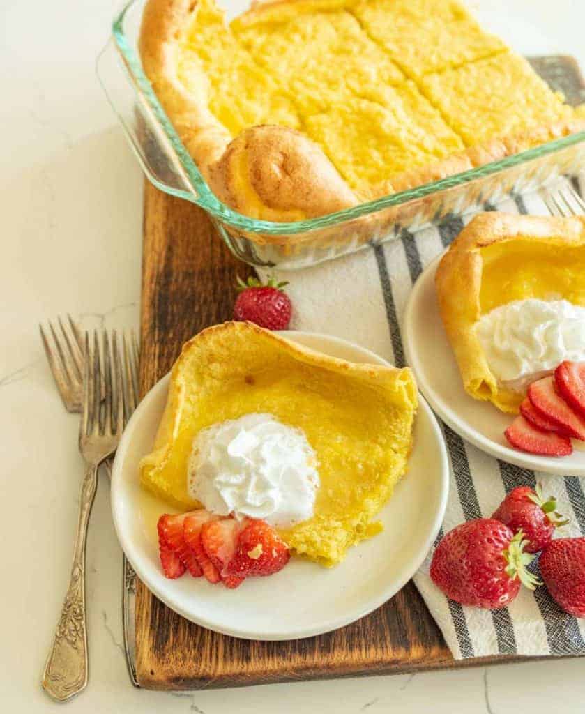 A plate with a serving of Dutch baby, also known as a German pancake recipe, is topped with whipped cream and sliced strawberries. It sits on a cutting board, with extra pancake in a baking dish and another plate in the background. Two forks rest nearby.