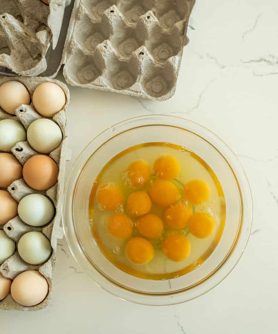 Top view of cracked raw eggs in a glass bowl next to a carton of different colored farm eggs.