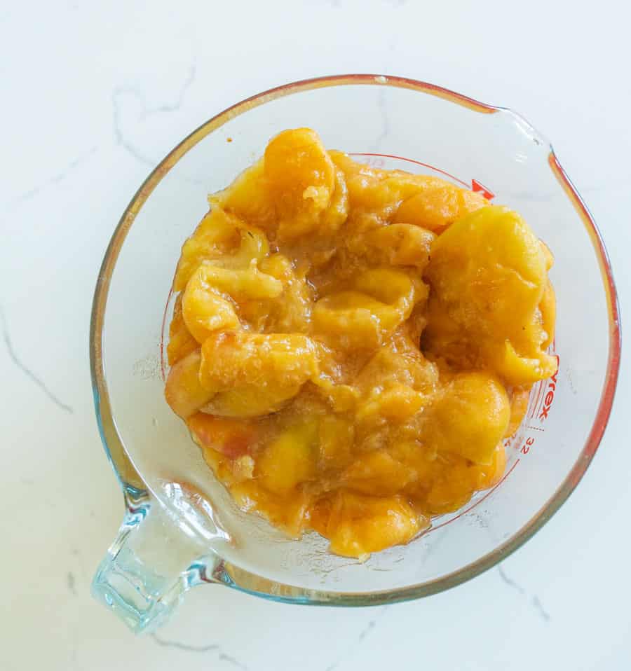 A glass measuring cup filled with mashed peaches sits on a white marble countertop. The peaches appear soft and saucy, with a golden-yellow color&mdash;perfect for making easy  homemade fruit leather snacks for kids.
