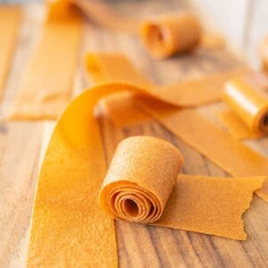 Wide strips of homemade fruit leather are rolled up and laid out on a wooden surface. Some pieces remain flat, while others are partially or fully rolled.