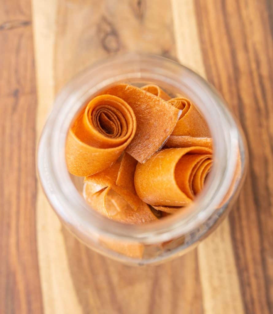 A top-down view of a glass jar filled with rolled homemade fruit leather strips, sitting on a wooden surface with light and dark grain patterns.
