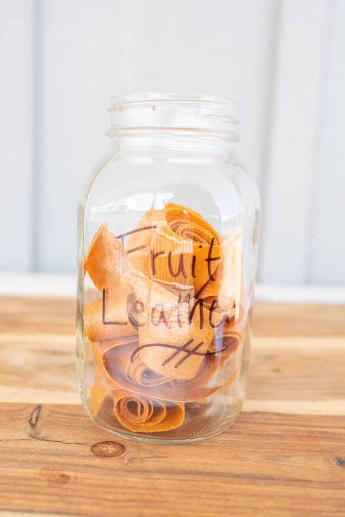 A glass jar labeled Fruit Leather contains several rolled pieces of homemade fruit leather, their orange-brown hues contrasting beautifully against the wooden surface and light-colored background.