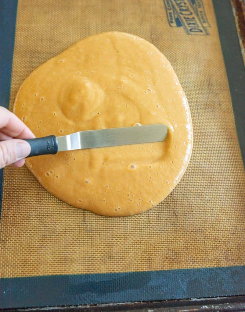 A hand uses an offset spatula to spread a thick, golden caramel mixture&mdash;similar in texture to homemade fruit leather&mdash;on a silicone baking mat placed on a baking sheet.