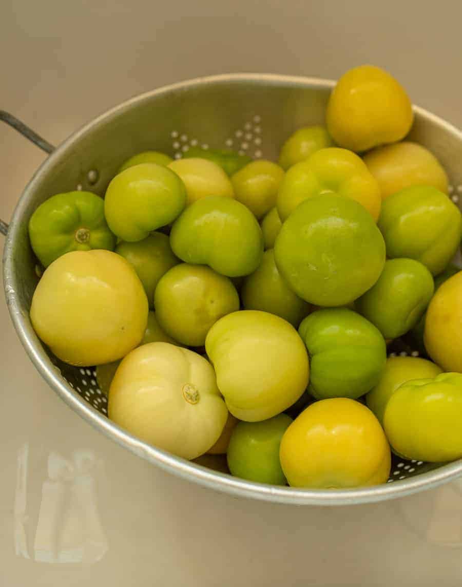 Colander filled with peeled tomatillos being washed in a white sink.