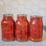 Three glass jars filled with canned tomatoes, preserved using the water bath method, are lined up on a white countertop. The jars have silver lids, and the background features a hexagonal tile pattern.