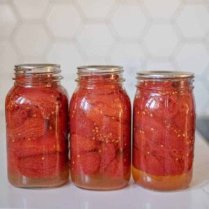 Three glass jars filled with canned tomatoes, preserved using the water bath method, are lined up on a white countertop. The jars have silver lids, and the background features a hexagonal tile pattern.