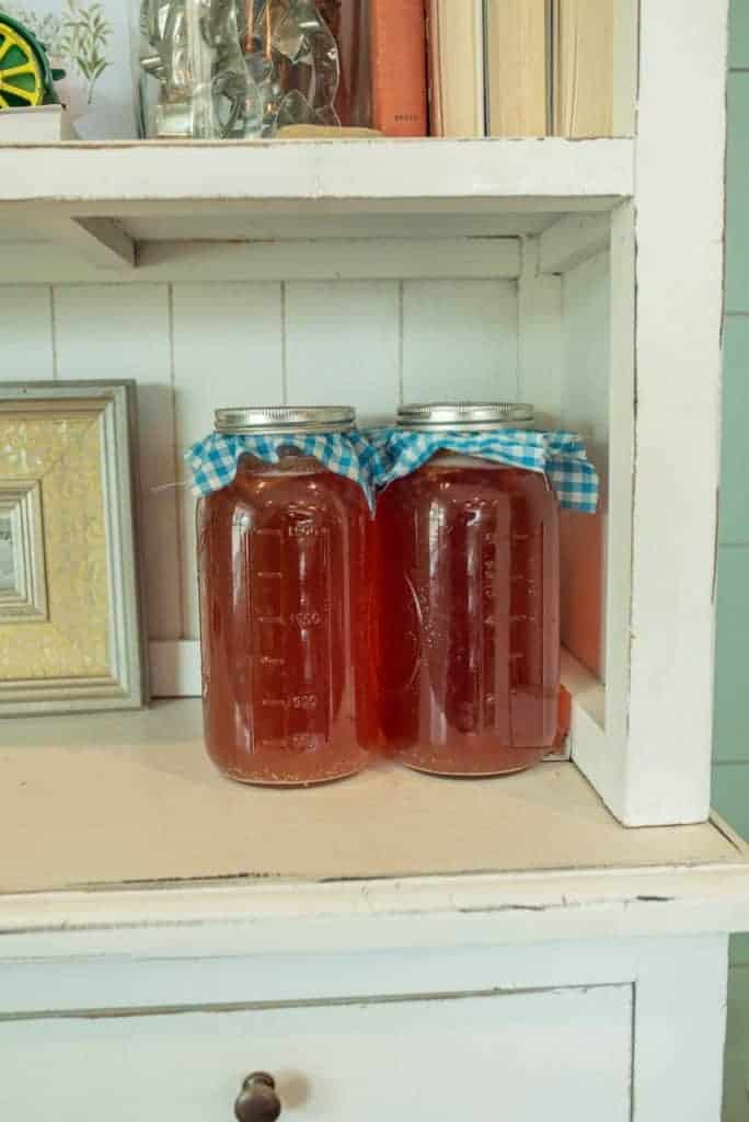 two half gallon mason jar filled with first ferment kombucha and capped with gingham cloth and metal ring sitting on pretty hutch.
