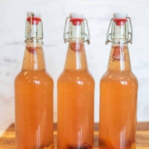Three clear glass bottles with swing-top lids are filled with a light orange, fizzy homemade kombucha. Pieces of fruit rest at the bottom of each bottle. The bottles sit on a wooden surface against a light background.