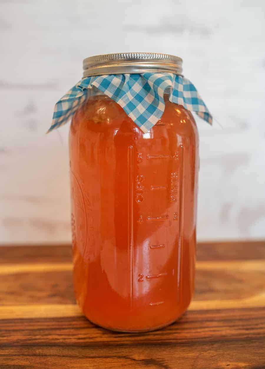 A jar of an orange colored liquid with a metal lid rests atop a blue checkered cloth on a wooden surface.