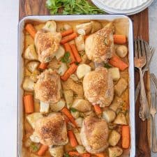 oven roasted chicken thighs, carrots, potatoes, and onions on enamel baking sheet with dinner plates, forks, and garnish