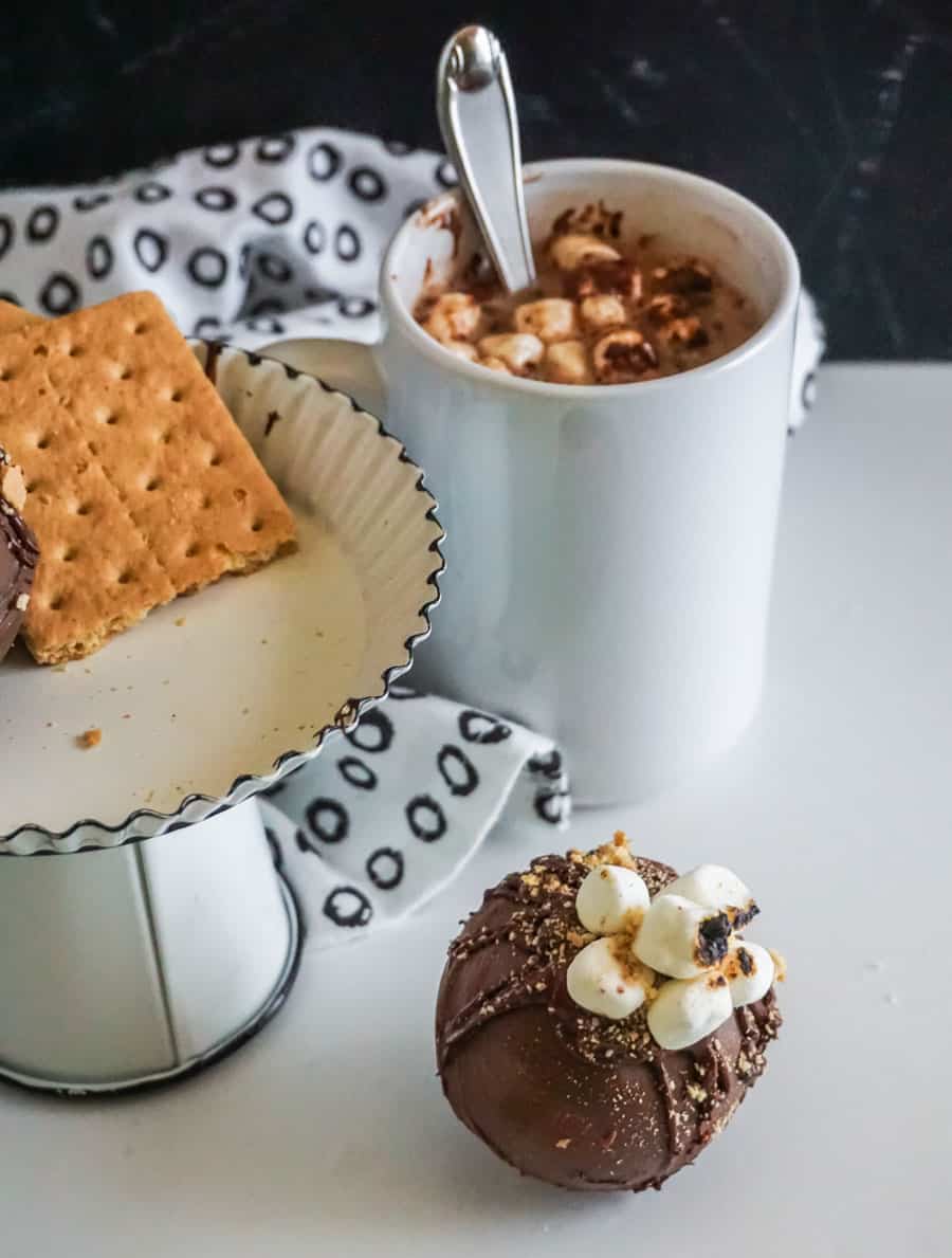 A hot chocolate bomb on a white counter next to a white enamel cake stand with graham crackers and bombs on it and a white mug with hot cocoa, marshmallows and a spoon.