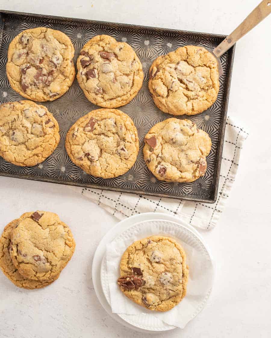 A giant chocolate chunk cookie on a white plate next to a baking sheet with six more cookies on it.