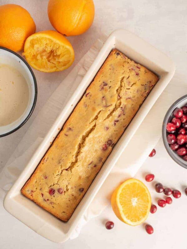 loaf of cranberry orange bread in white loaf pan surrounded by oranges and cranberries