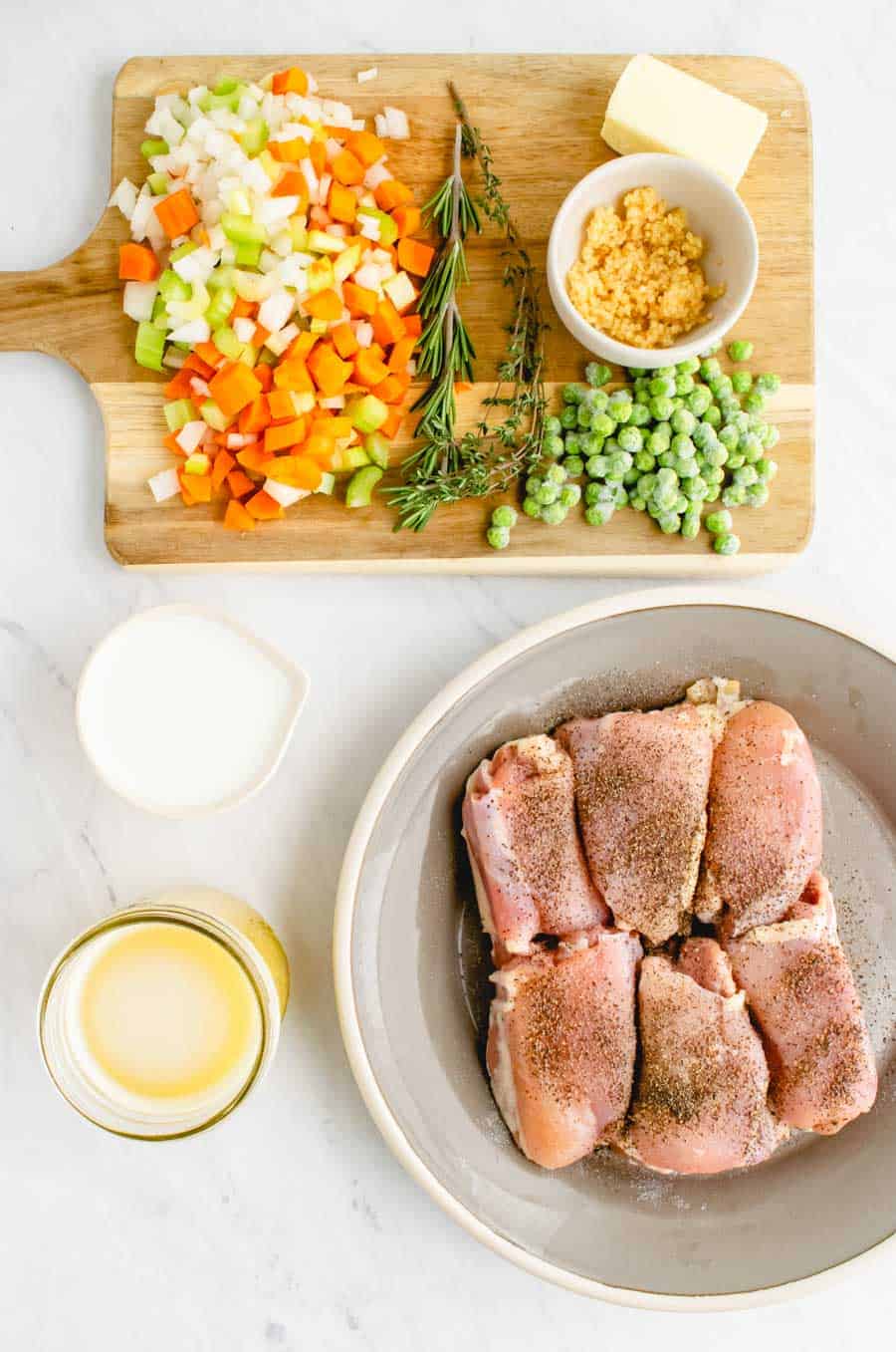 Ingredients to make chicken stew with dumplings including raw chicken thighs on a grey plate and peas, herbs, carrots, celery, onions, and garlic on a wooden cutting board.