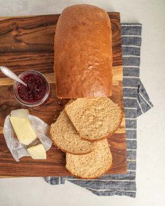 A loaf of whole wheat sandwich bread with three slices cut, a jar of red jam with a spoon, and pieces of butter on a wrapper are arranged on a wooden cutting board over a striped cloth.