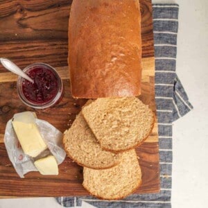 A loaf of whole wheat sandwich bread with three slices cut, a jar of red jam with a spoon, and pieces of butter on a wrapper are arranged on a wooden cutting board over a striped cloth.