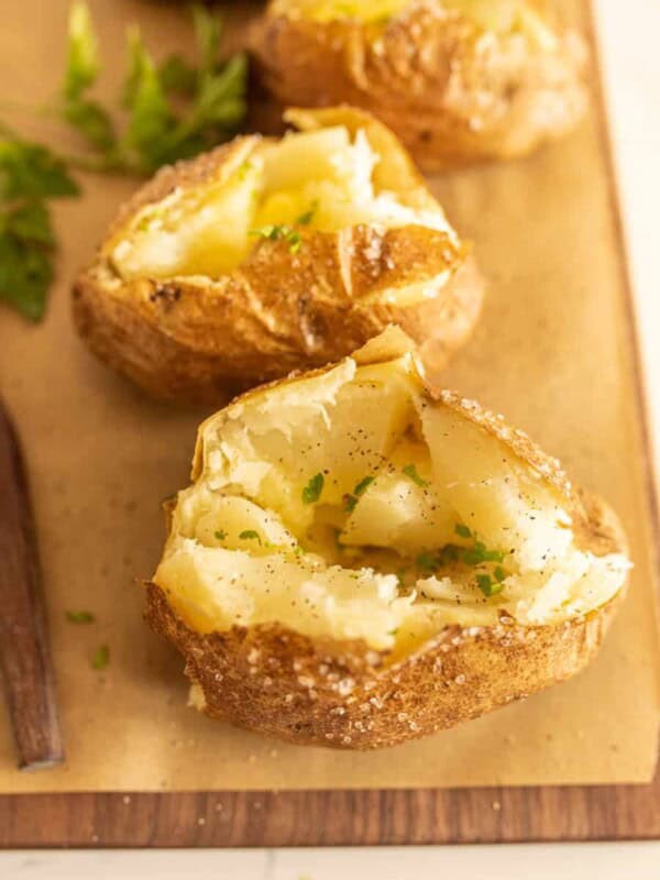An oven baked potato split open, topped with melting butter, black pepper, and chopped parsley, sits on parchment paper with more oven baked potatoes and fresh parsley in the background.