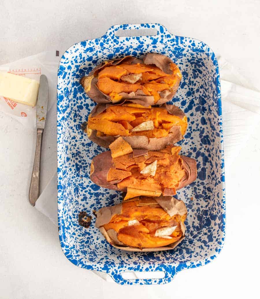 Top view of baked sweet potatoes in a white and blue speckled baking dish next to a butter knife and stick of butter.