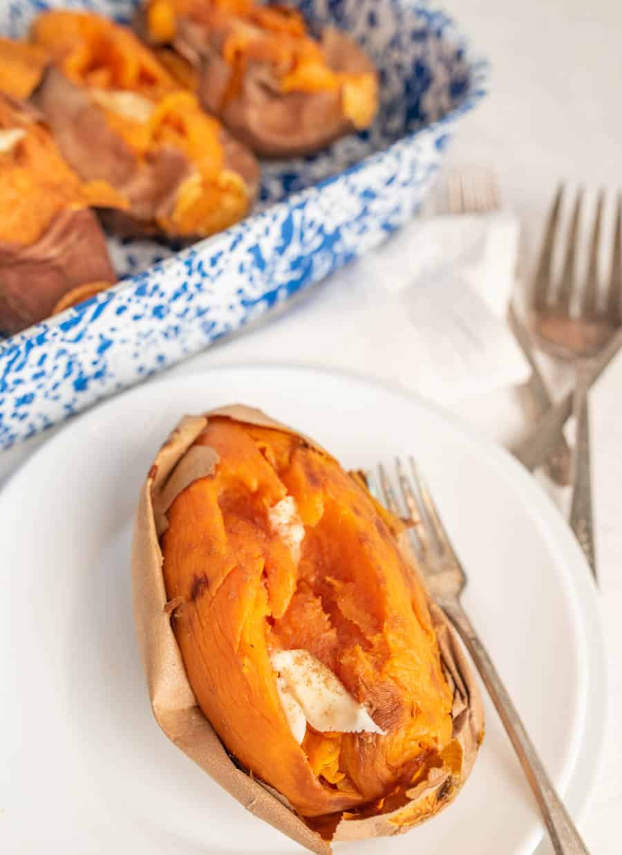 A baked sweet potato with butter on it on a white plate with a fork and three baked sweet potatoes in the background in a white and blue speckled baking dish.